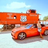 Adèle en robe est adossée à une Chevrolet Camaro SS rouge, devant un locomotive rouge avec Route 66 indiqué dessus.