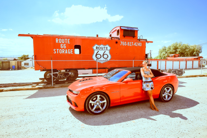 Adèle en robe est adossée à une Chevrolet Camaro SS rouge, devant un locomotive rouge avec Route 66 indiqué dessus.