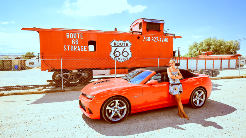 Adèle en robe est adossée à une Chevrolet Camaro SS rouge, devant un locomotive rouge avec Route 66 indiqué dessus.
